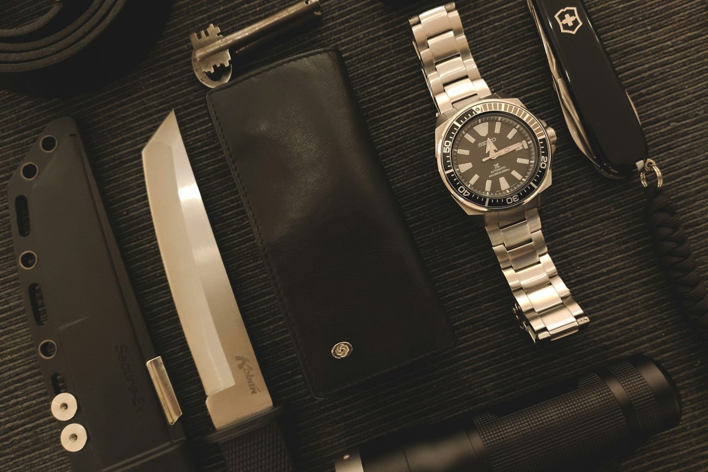 Clean flat lay of everyday carry gear including a flashlight, multitool, folding knife, notebook, and compass